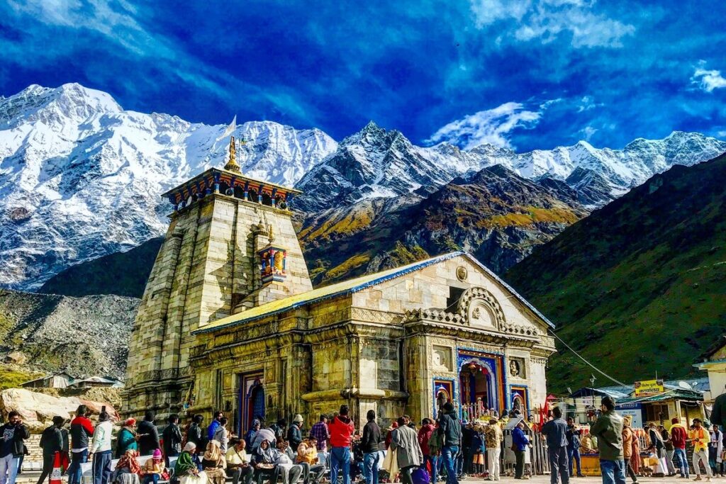 Kedarnath Temple with a backdrop of snow-covered peaks, with pilgrims walking towards the shrine.